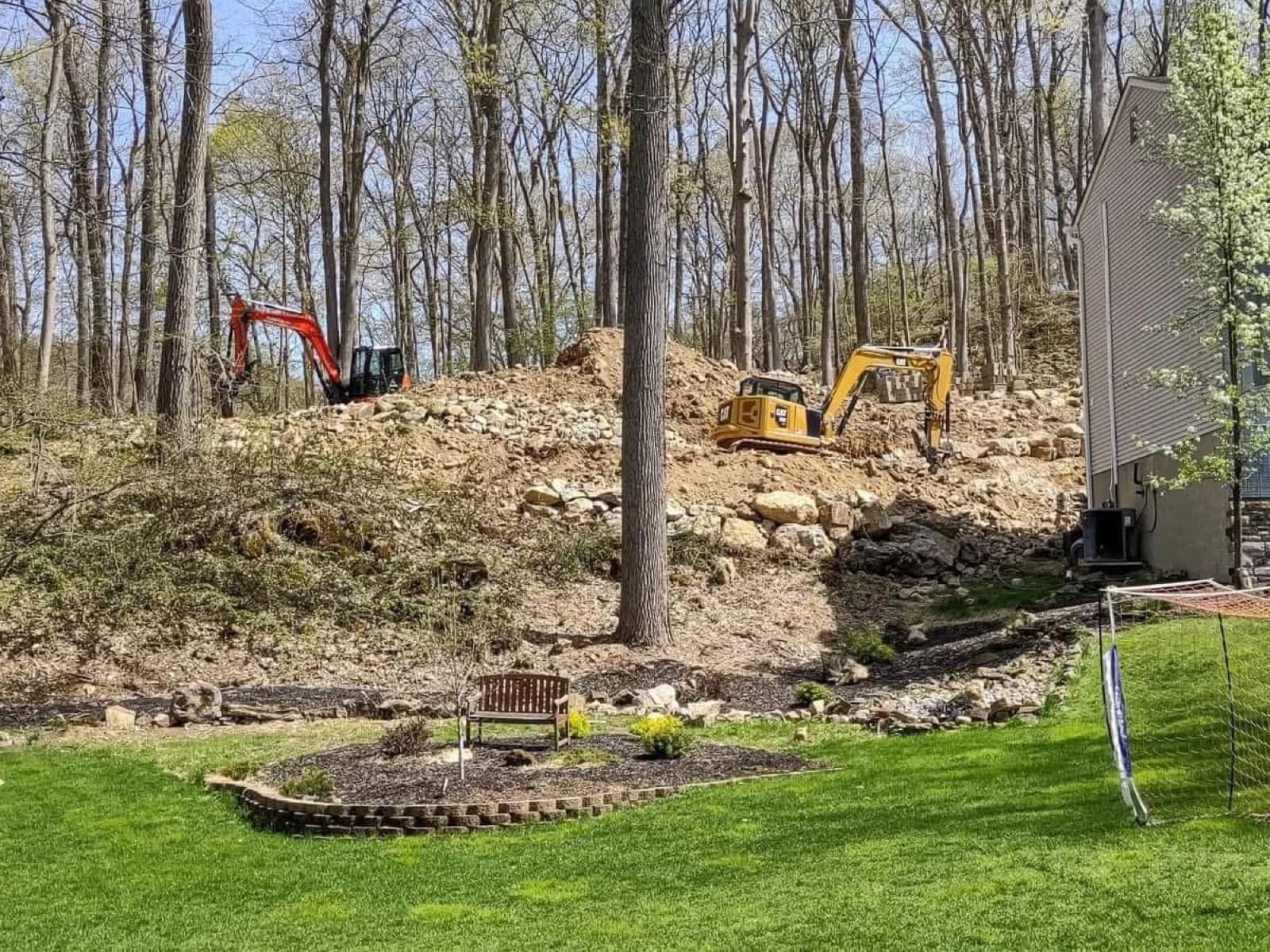 Excavators building a boulder retaining wall into a backyard hillside above a lawn
