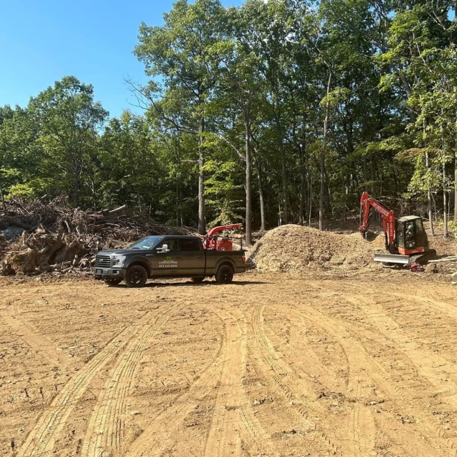 Freshly graded lot with crew truck, wood chipper, and mini excavator at a wooded clearing site