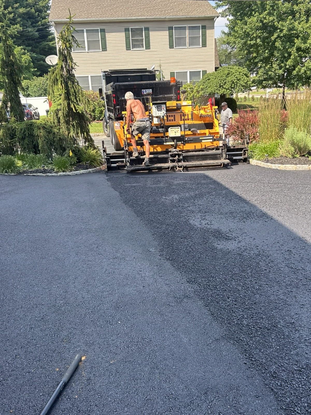 Crew running an asphalt paver fed by a dump truck across a residential driveway