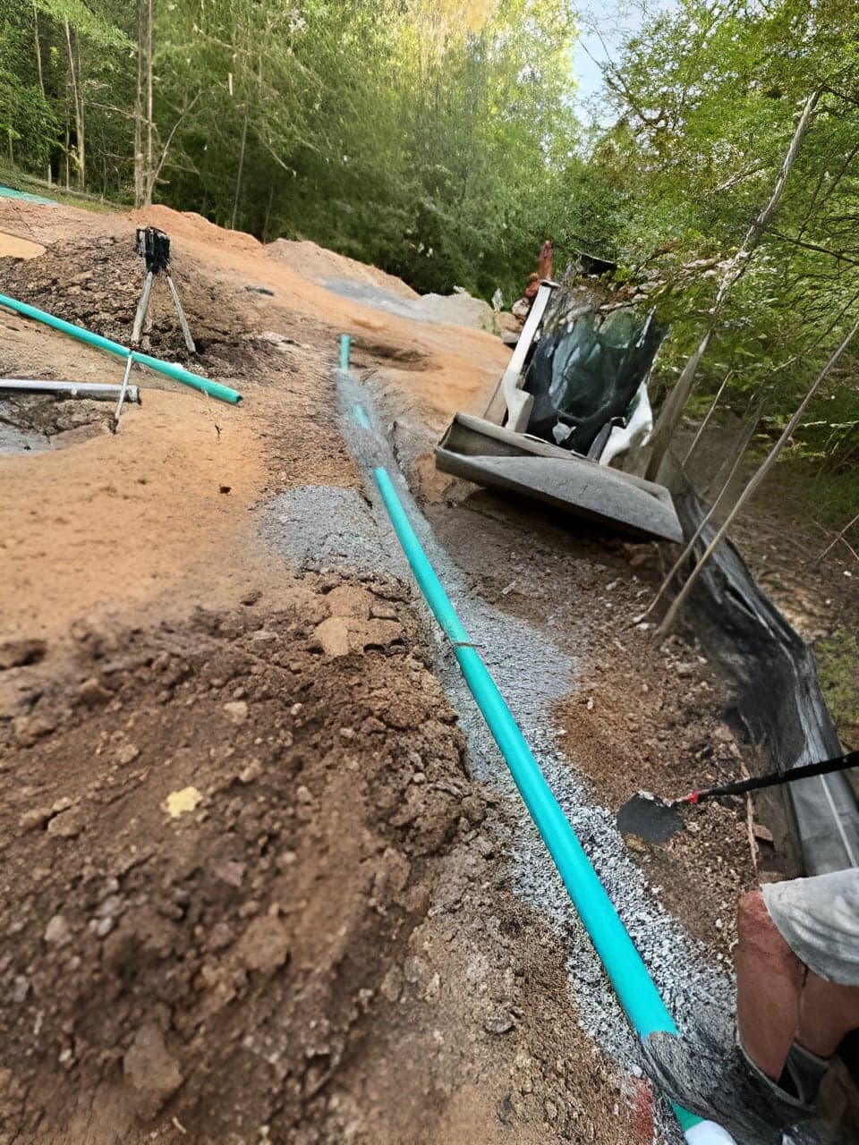 Long green drainage pipe run heading downhill past a skid steer on a wooded site