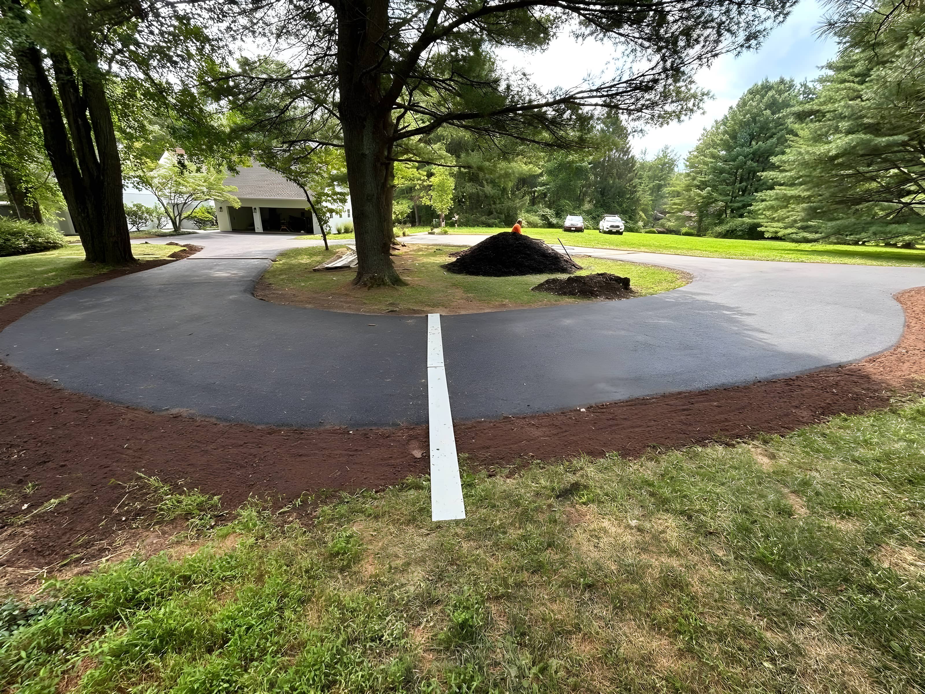 Wide view of a just-paved loop driveway with topsoil-finished edges around a tree island