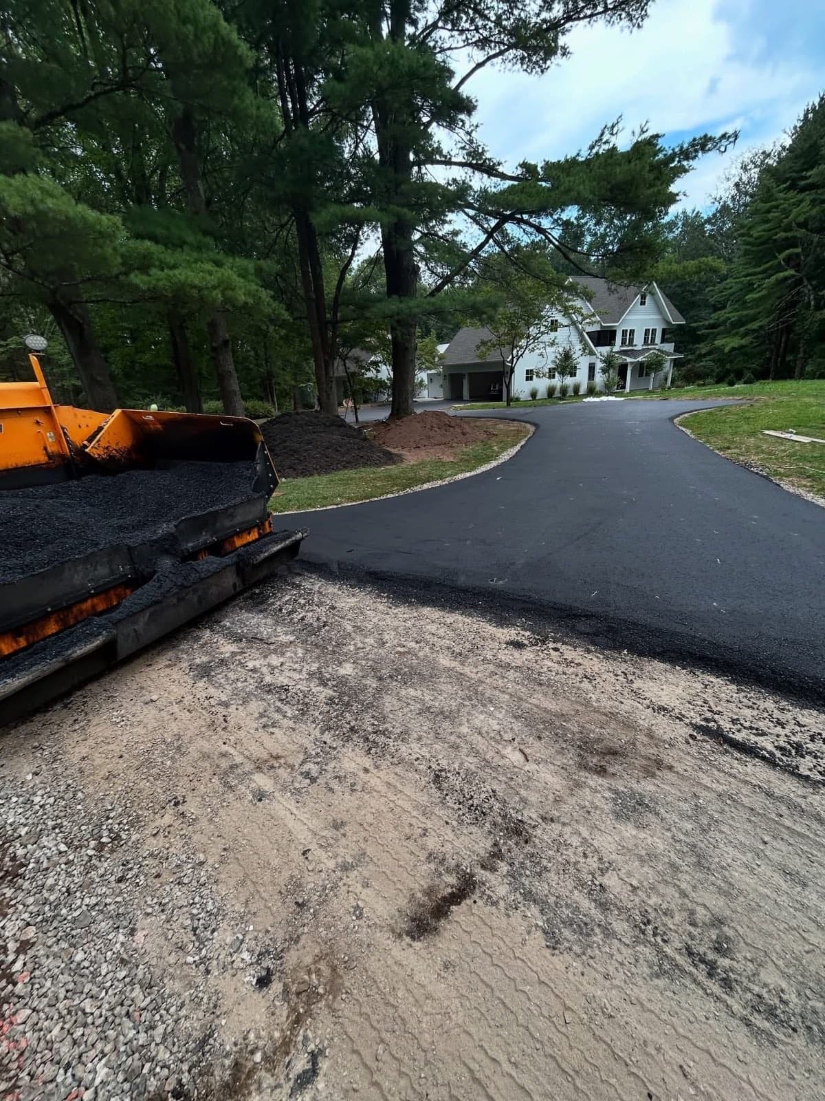 Paver hopper loaded with hot asphalt and a freshly paved driveway sweeping to a farmhouse