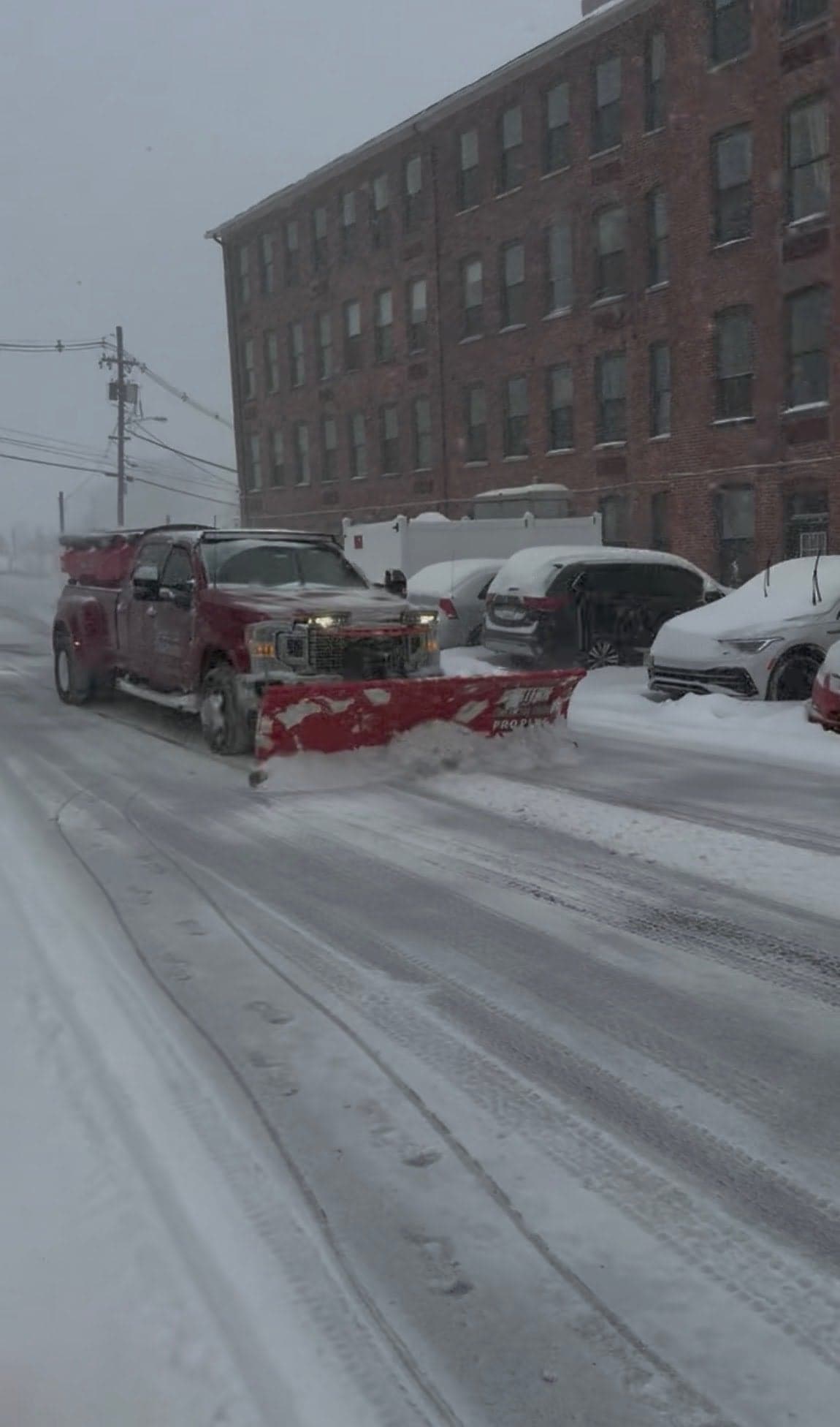 Plow truck clearing a snow covered access road past parked cars in a storm