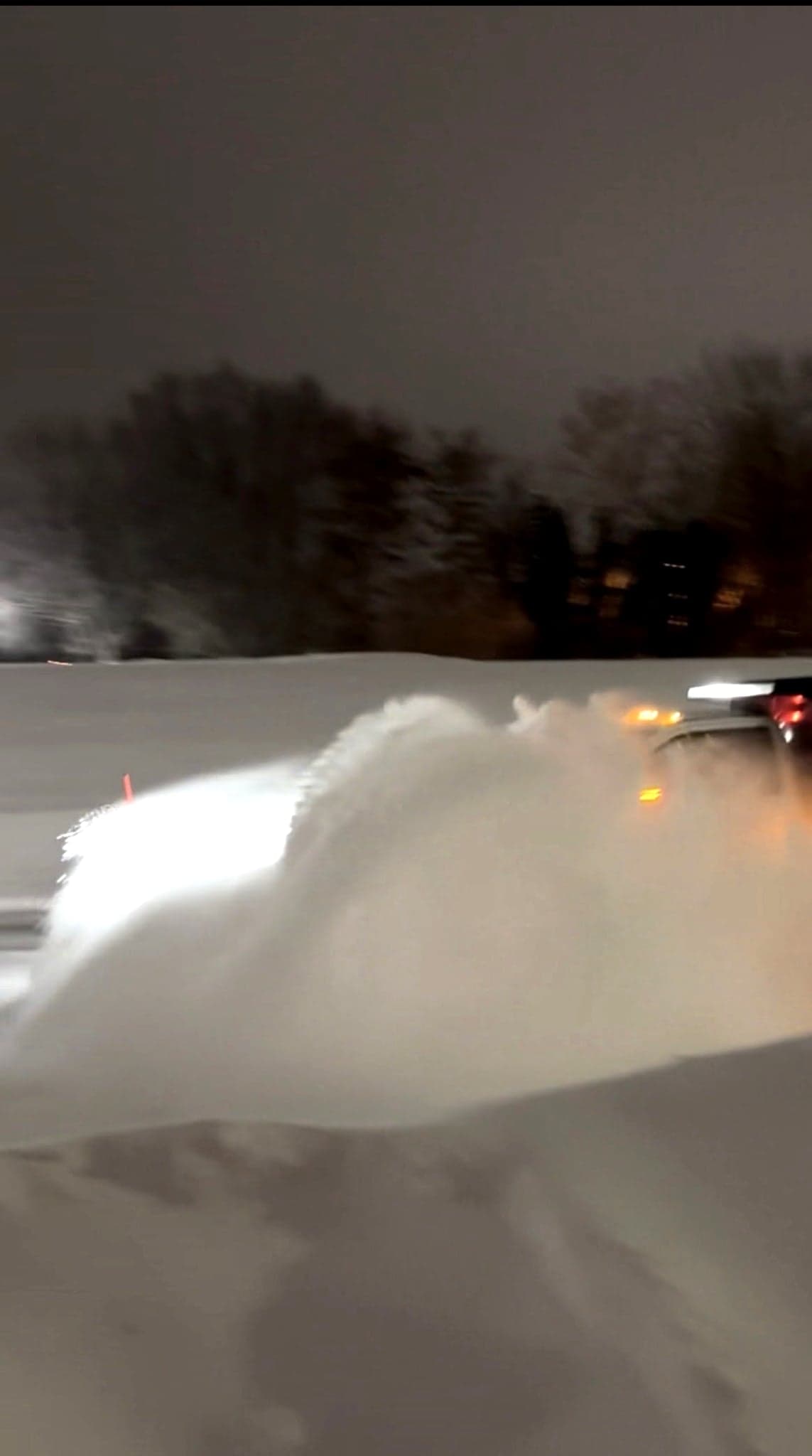 Plow blade throwing a wave of snow across a lot at night