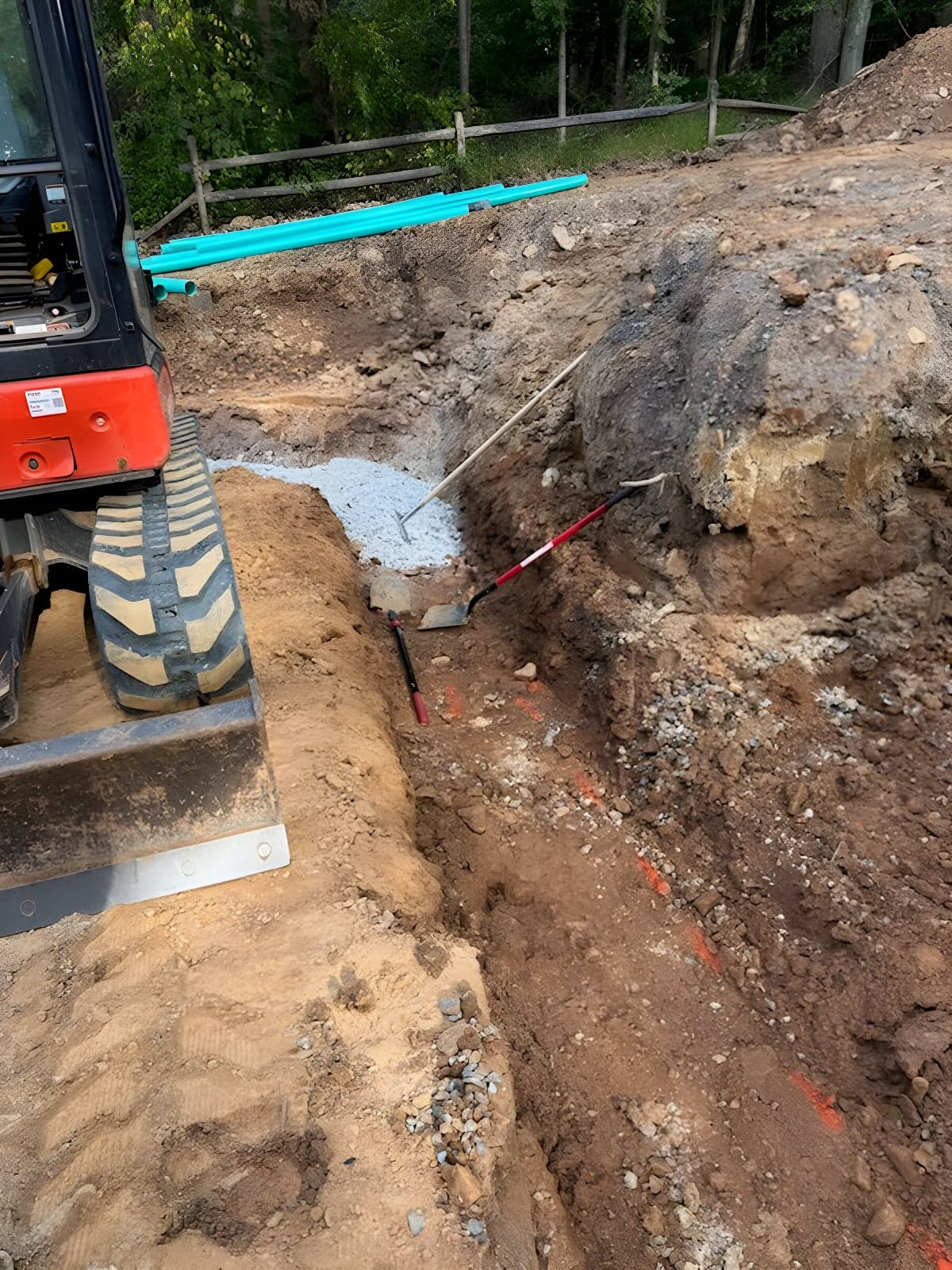 Excavator at an open trench with a gravel pocket and green pipe staged behind